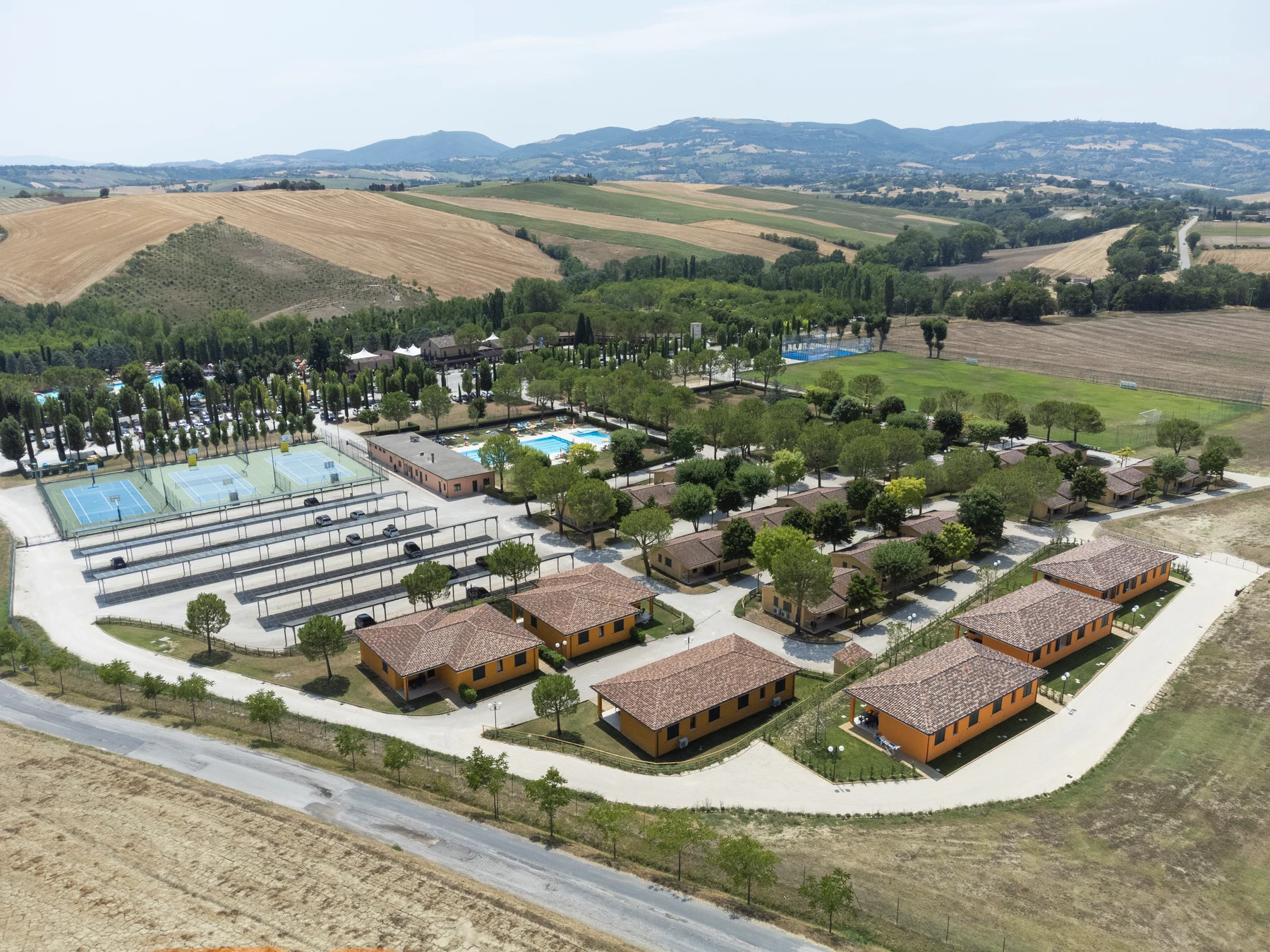 At Centro Vacanze Verde Azzurro, a bird's-eye perspective reveals cabins positioned around athletic amenities, encircled by meadows and far-off mountains and hills.