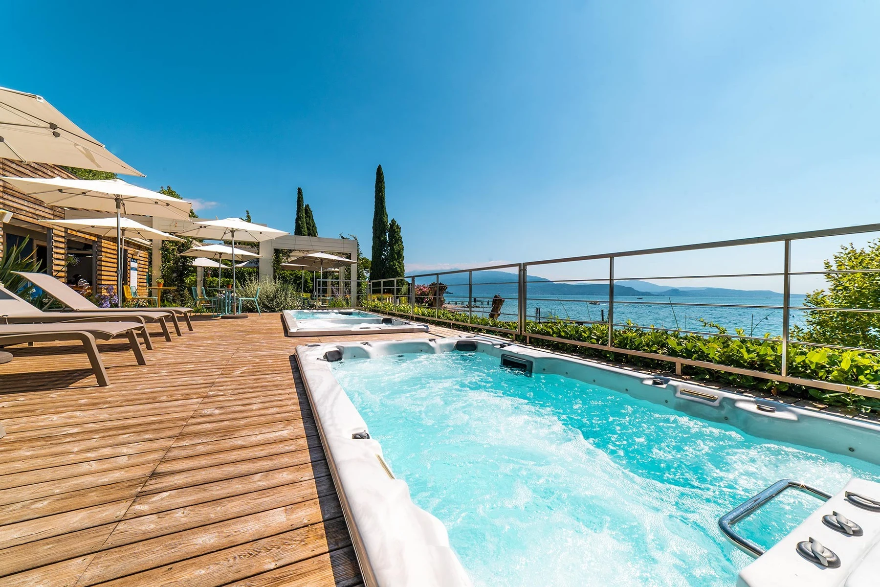 At La Gardiola, a hot tub froths on a timber terrace, surrounded by sunbeds and parasols, providing a picturesque view of a tranquil lake.