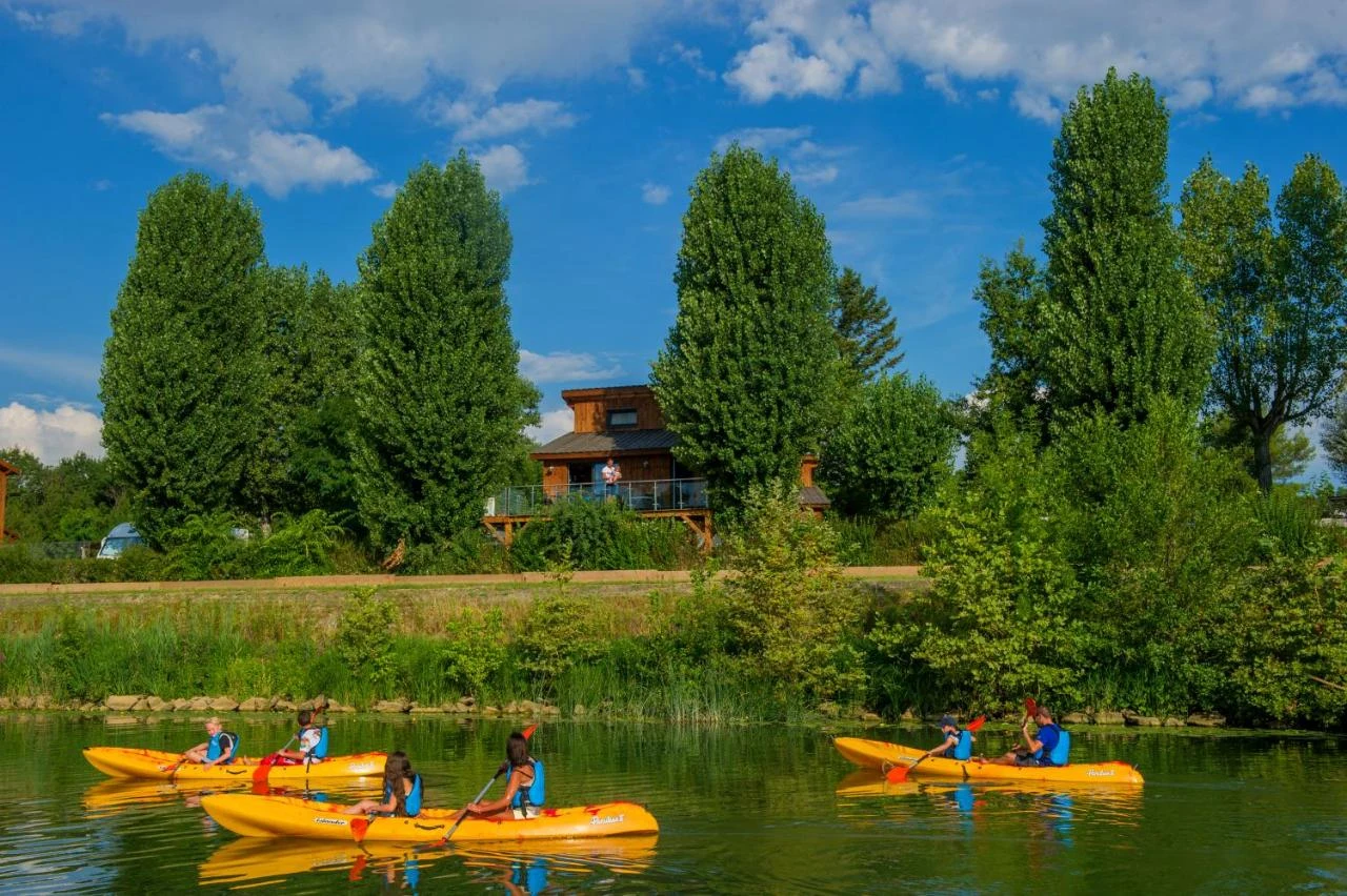 At Kanopée Camping & Lodge, kayakers navigate a lake encircled by vibrant foliage and trees, with a charming cabin in the backdrop.