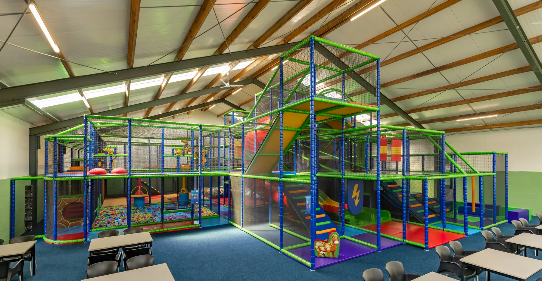 At Vakantiepark Breebronne, there is an indoor play area featuring a climbing structure with slides and a ball pit, located next to tables and chairs, all within a large room.