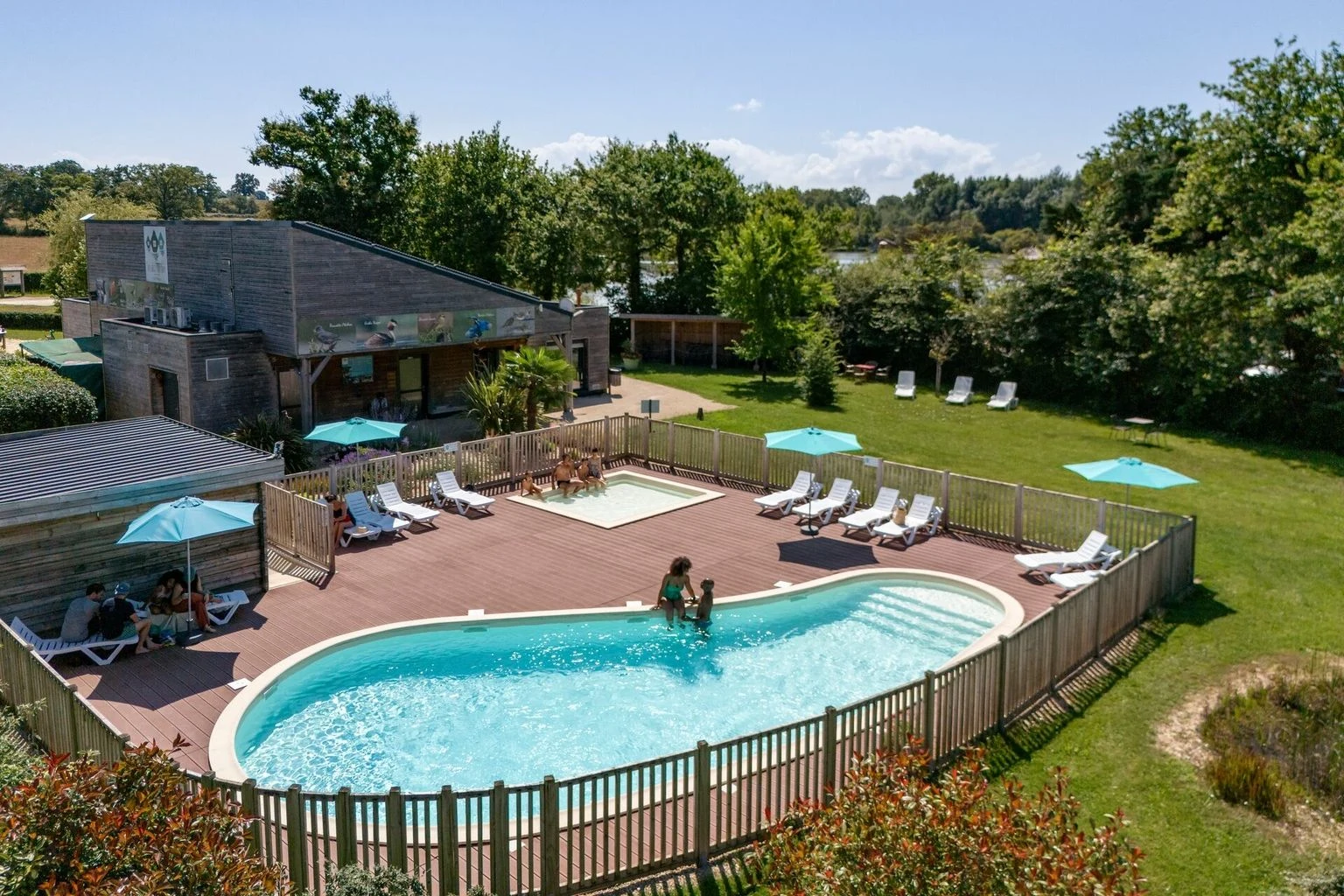 At Village Flottant de Pressac, a swimming pool with bathers is encircled by wooden decking, sun loungers, and parasols, situated in a verdant campsite.