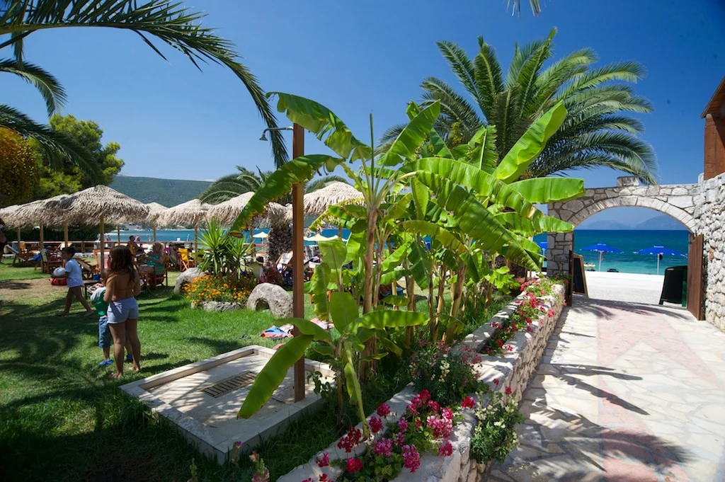 At Karavomilos Beach, individuals are unwinding and strolling near a seaside café adorned with palm trees and umbrellas, with a view of the ocean.