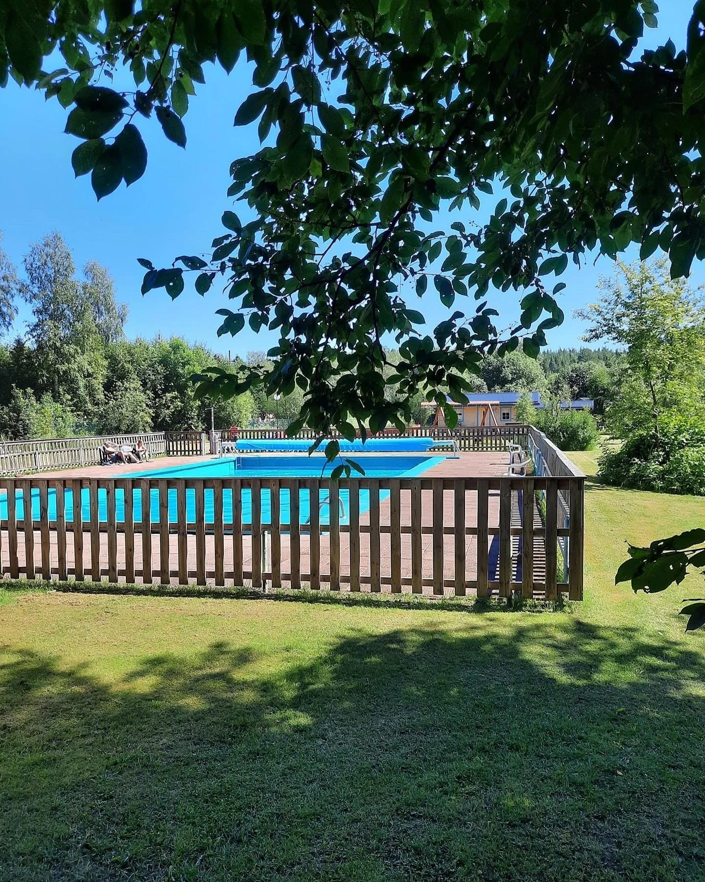 At Yttermalungs, a swimming pool enclosed by a wooden barrier is nestled among trees and grass, with individuals lounging in the sun nearby.