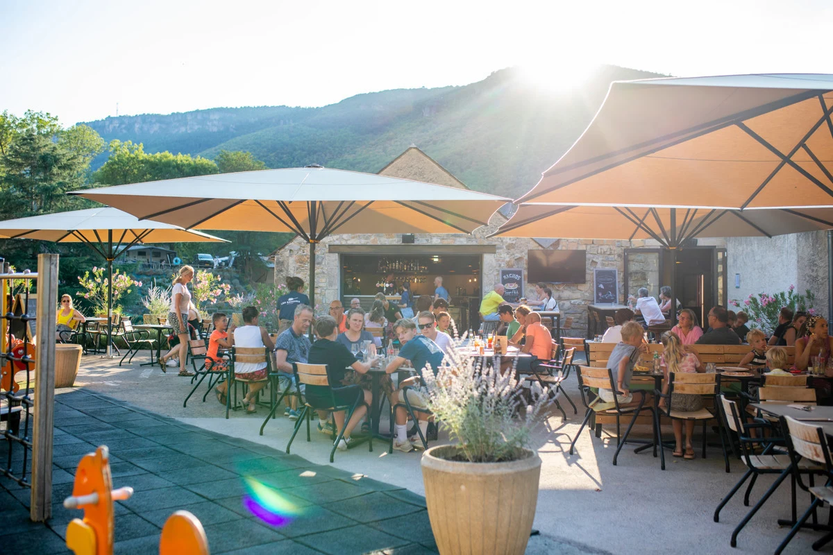 At Val de Cantobre, individuals are enjoying meals outside beneath large canopies, surrounded by a mountainous scenery on a bright day.