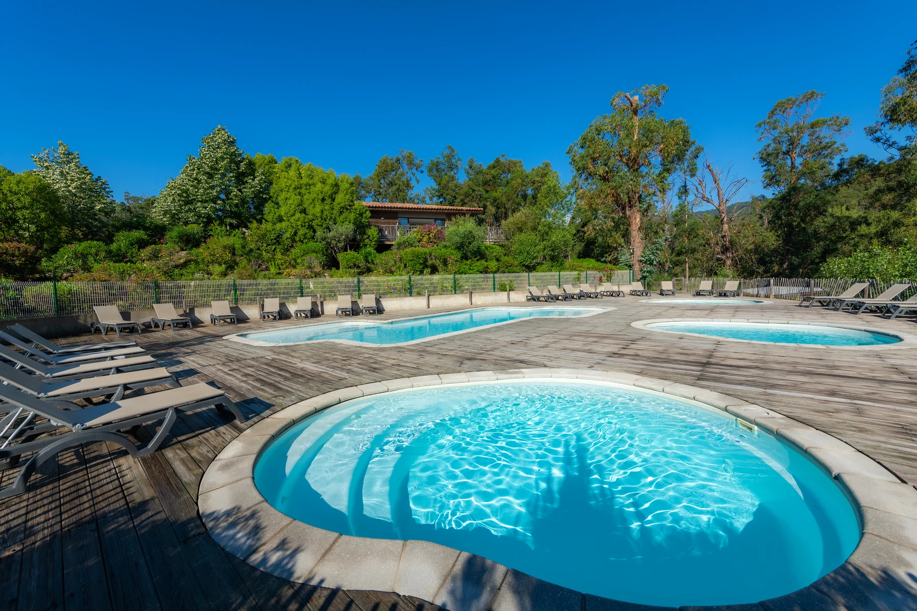 At Sole di Sari, circular and rectangular swimming pools mirror sunlight, encircled by lounge chairs on wooden decking, amidst vibrant green vegetation.