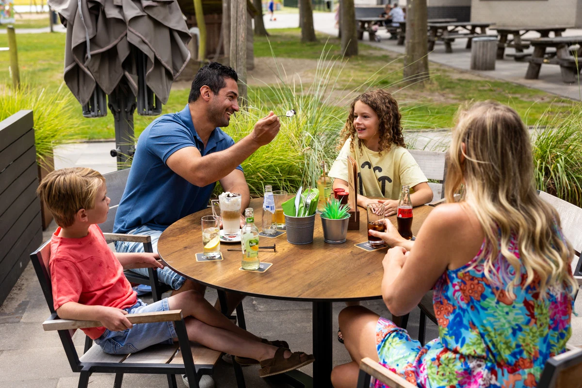 At de Flaasbloem, four individuals are gathered around a circular outdoor table, enjoying beverages and sweets in a lush, tree-filled park.