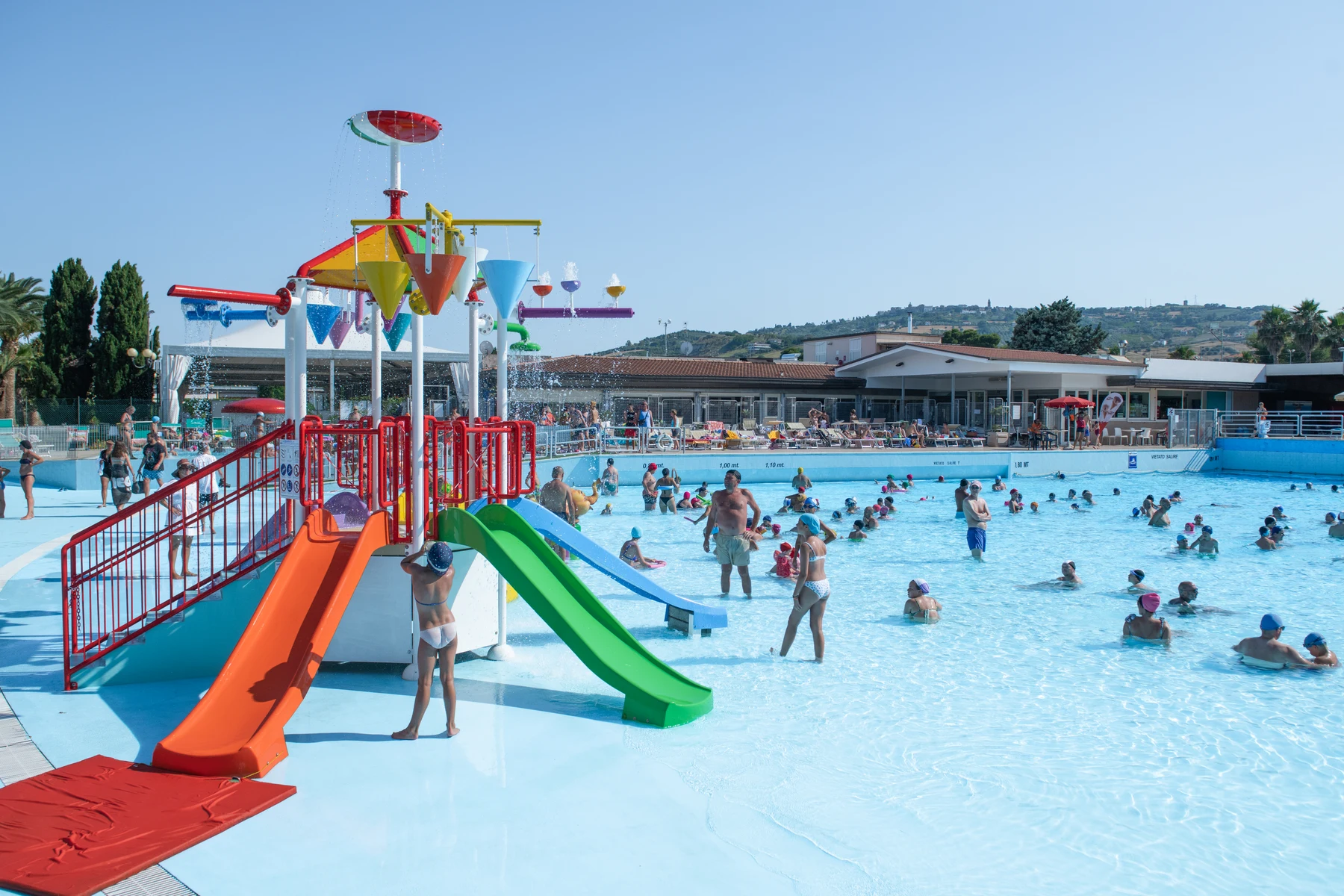 At Villaggio Turistico Lido D'Abruzzo, a vibrant water play area featuring slides delights children, encircled by individuals swimming in a spacious outdoor pool.
