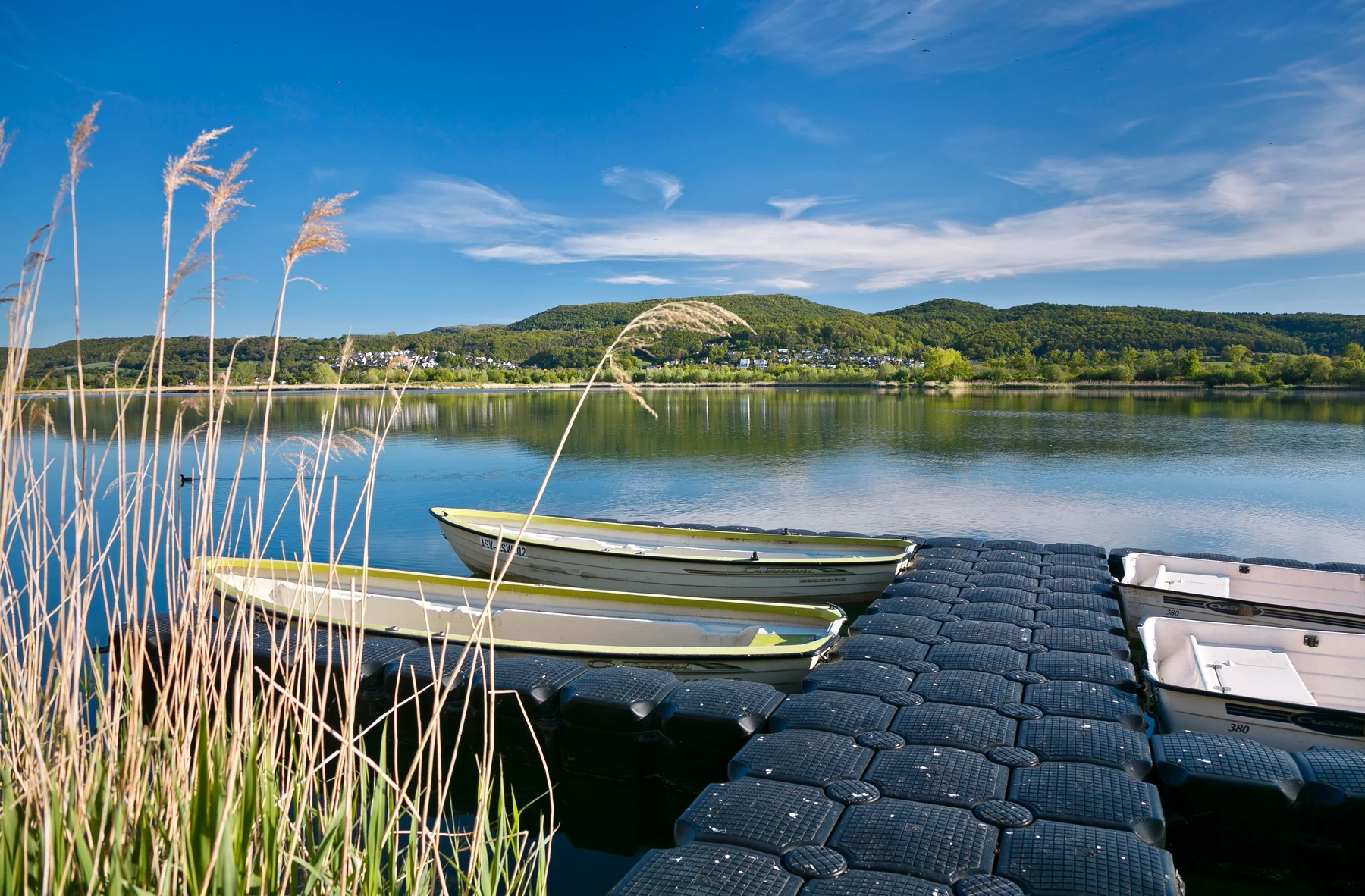 At KNAUS Campingpark Eschwege, rowboats moored at a buoyant dock on a tranquil lake, encircled by hills and reeds.