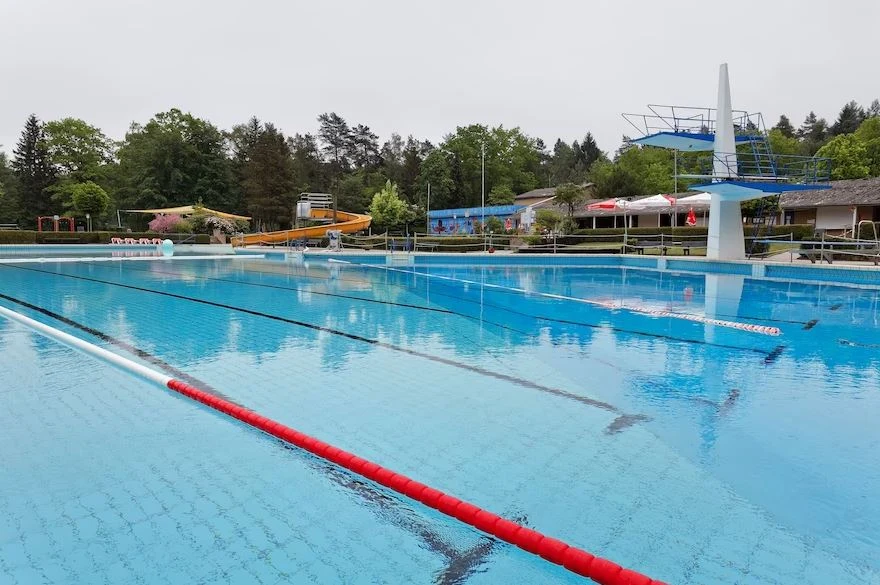 At Knaus Campingpark Bleckede/Elbtalaue, there is an outdoor swimming pool featuring diving boards, lane indicators, and a water slide in the backdrop, encircled by trees and structures.