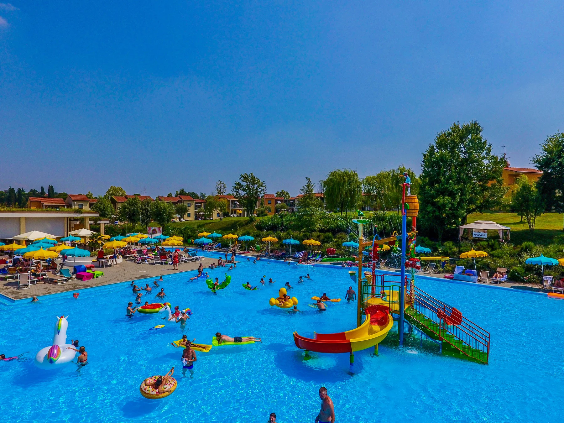At Belvedere Village, a vibrant pool features bright slides and inflatable floats; individuals are swimming and unwinding, encircled by trees.