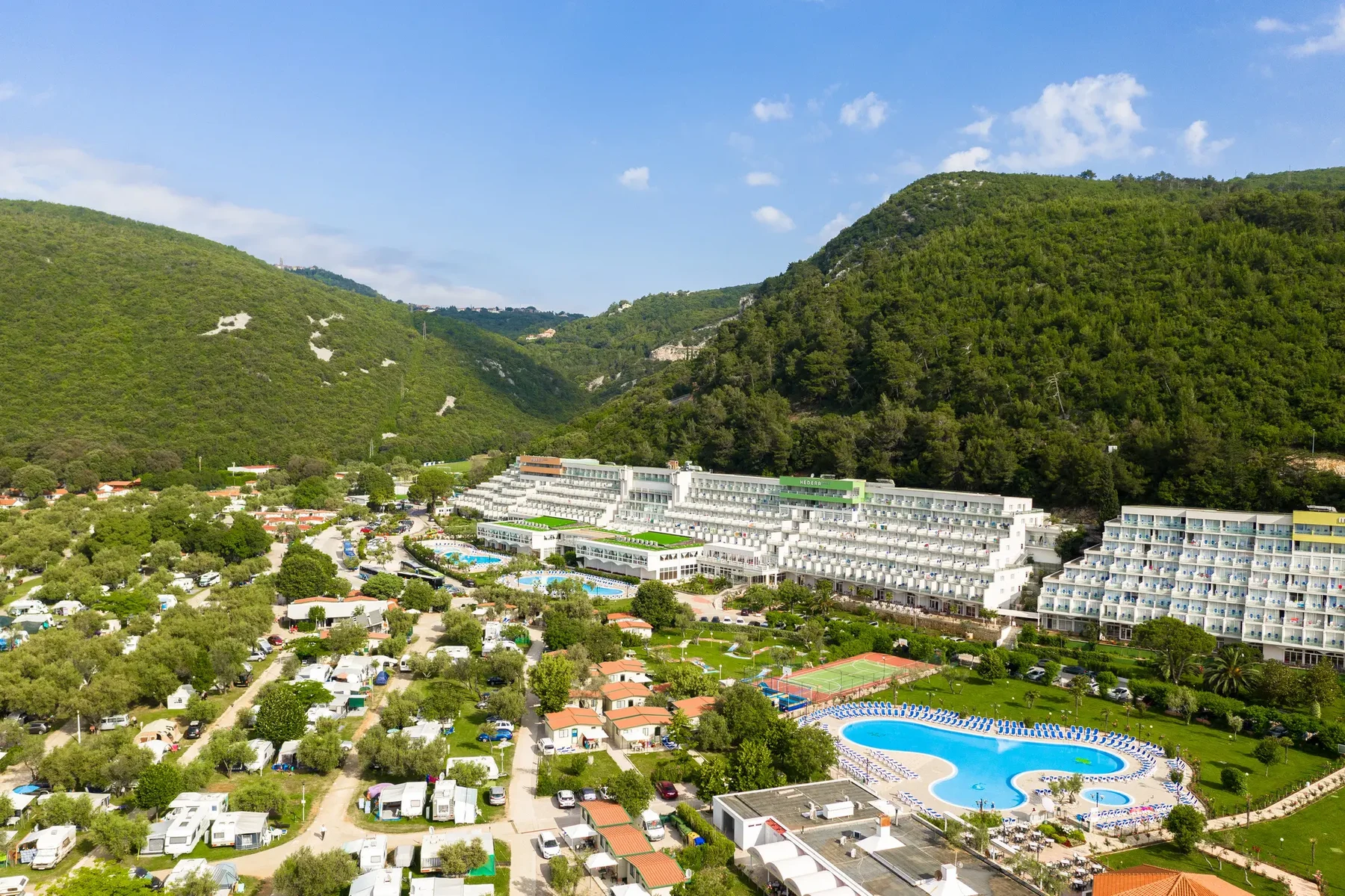 At Oliva, a hotel complex overlooks swimming pools, nestled among verdant hills and a campsite featuring caravans and tents.