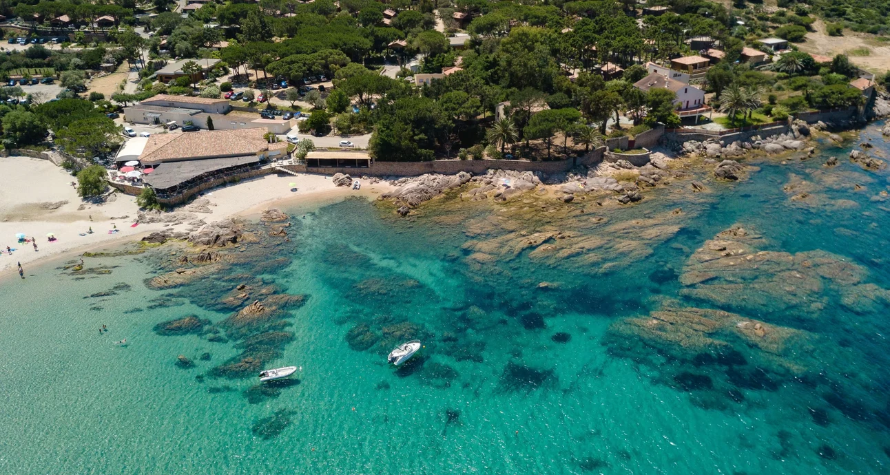 At U Livanti, vessels drift on transparent turquoise waters close to a rugged coastline and sandy shore, encircled by trees and structures.