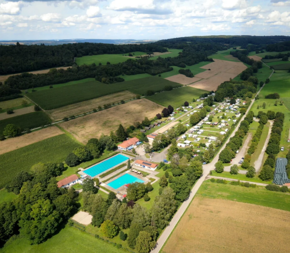 At Walsheim, an aerial perspective reveals a campsite featuring swimming pools, encircled by meadows and wooded hills.