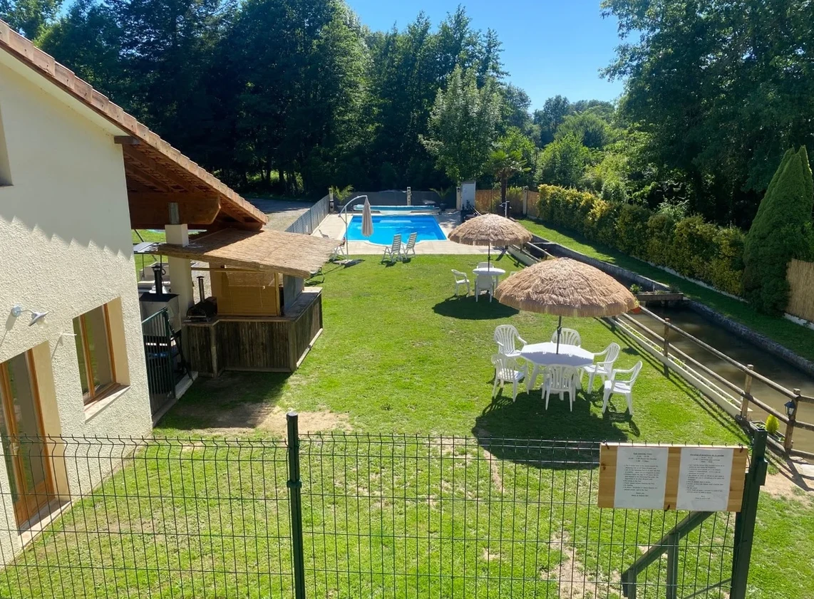 At Moulin du Pommier, there is a swimming pool accompanied by lounge chairs and umbrellas situated on a grassy lawn adjacent to a structure, encircled by trees.