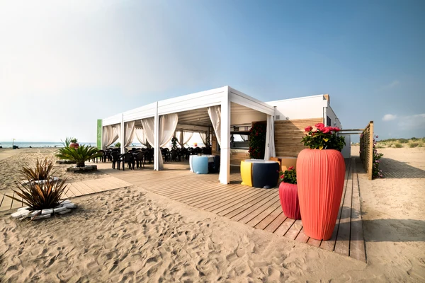 At Marina di Venezia, there is a pavilion featuring potted greenery and seating spaces on a wooden deck adjacent to the sandy shore.