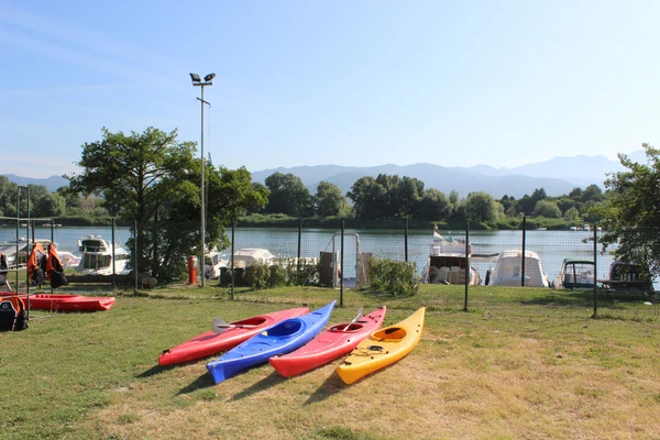 At River, vibrant kayaks are situated on the grass beside a lake, accompanied by anchored boats and distant hills in the background.