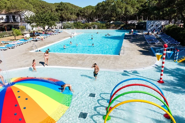 At Neptuno, individuals are enjoying themselves in a shallow wading pool adorned with vibrant arches, with a spacious pool in the background, encircled by pine trees.