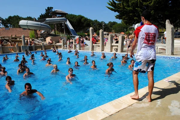 At Le Vieux Port, an individual is conducting a water aerobics session in an outdoor swimming pool featuring a large water slide and trees in the background.
