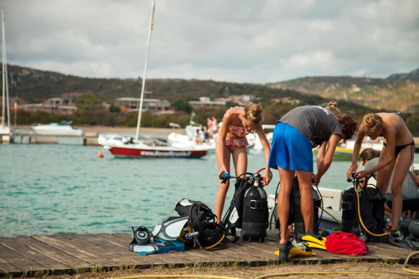 At Isuledda, individuals are assembling scuba diving equipment on a pier next to serene, turquoise waters, with boats and distant hills in the background.