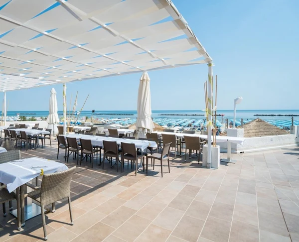 At the outdoor dining establishment, adorned with wicker furnishings and sheltered by a pergola, guests can enjoy views of a beach dotted with umbrellas and the tranquil sea in Tahiti.