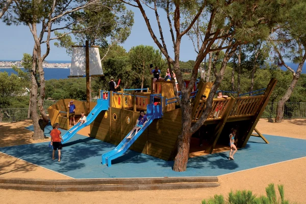 At Internacional de Calonge, children enjoy playing on a ship-inspired playground surrounded by trees, offering a picturesque view of the water and a distant city skyline.