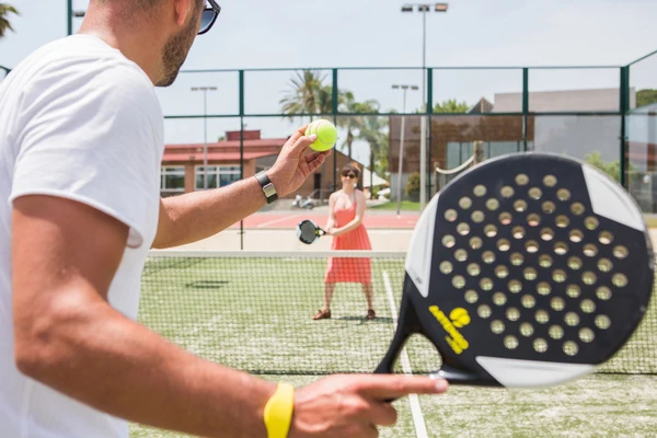 At Cambrils Park, two individuals are engaged in paddle tennis, with one player serving a ball to the other over a net on an outdoor court.