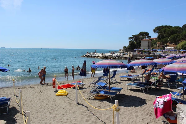 At River, sunbathers unwind beneath parasols and bathe in the ocean at a bustling sandy shore, with rocky formations and structures in the vicinity.