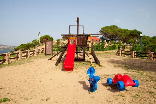 At Isuledda, there is a play area featuring a slide and a climbing wall situated on sandy terrain, encircled by trees and a wooden fence.