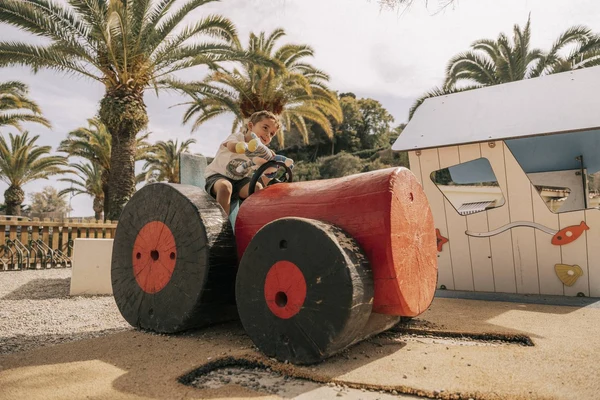 At Costa del Sol Glamping Village, a child rides a wooden tractor in a play area encircled by palm trees and recreational structures.