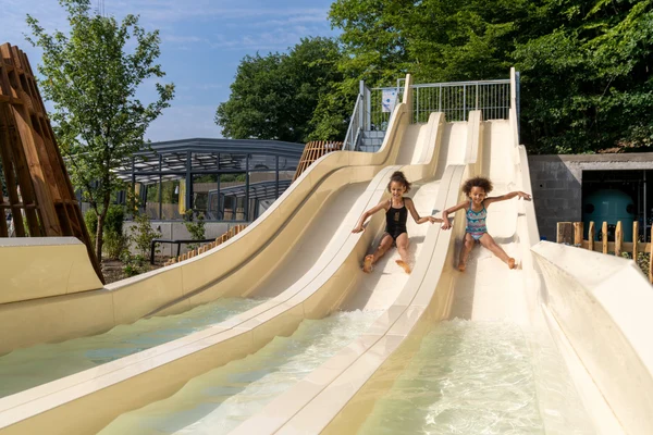 At Parc La Clusure, two kids glide down neighboring water slides, encircled by trees and outdoor facilities.