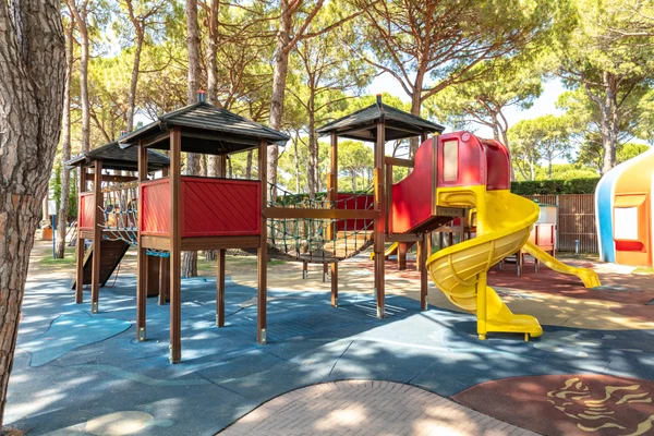 At Marina di Venezia, playground equipment featuring slides and rope bridges is situated beneath towering pine trees, enveloped by a natural setting.
