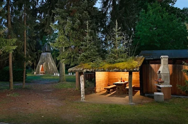 At Knaus Campingpark Bleckede/Elbtalaue, a wooden lodge features a roof adorned with moss, an illuminated table within, and is encircled by towering trees; a tipi and stone barbecue are located nearby.