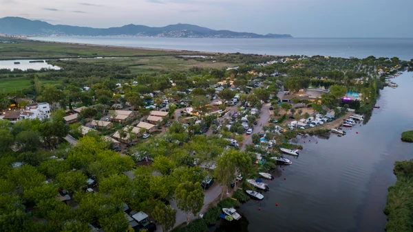 At Nautic Almata, tents and caravans are dispersed among trees, adjacent to a river with moored boats, providing a view of distant mountains and coastline.