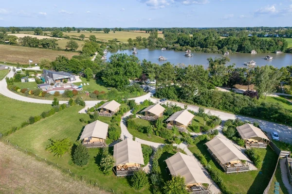 At Village Flottant de Pressac, safari-style tents are positioned in a lush, landscaped campground adjacent to a lake, featuring small, lakeside cabins nestled among the trees.