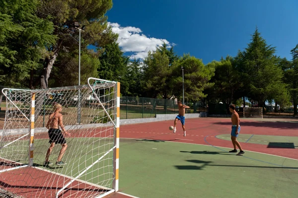 At Naturist Park Koversada, three shirtless individuals engage in soccer on an outdoor field encircled by trees.