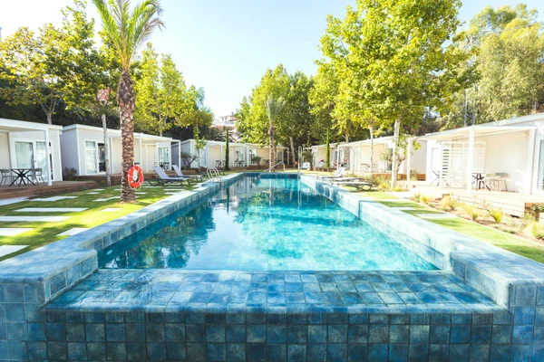 At Costa del Sol Glamping Village, a turquoise swimming pool is encircled by lounge chairs and small white cabins, nestled among vibrant green trees.
