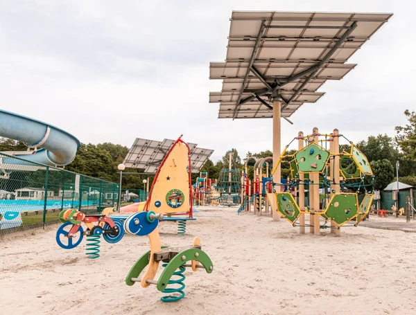 At Recreatieoord Wilhelm Tell, playground apparatus remains unused beneath solar panels on a sandy surface, accompanied by a nearby waterslide and enclosed swimming pool area.
