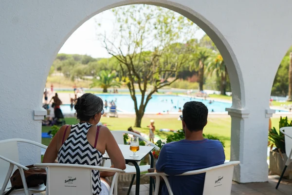 At Internacional Palamos, two individuals are seated beneath an arch, observing a bustling pool encircled by lush vegetation.