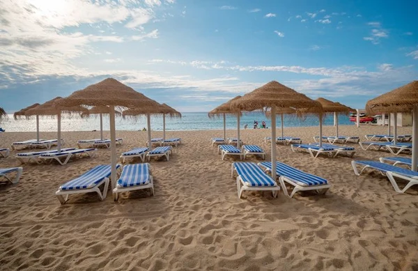 At Internacional de Calonge, sunbeds under thatched umbrellas are arranged on the sandy shore, facing the ocean.
