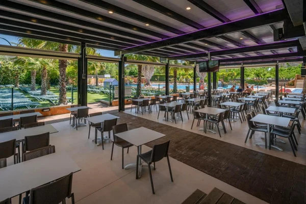 At Solmar, the al fresco dining space is adorned with tables and chairs, providing a view of a sunlit swimming pool and palm trees.