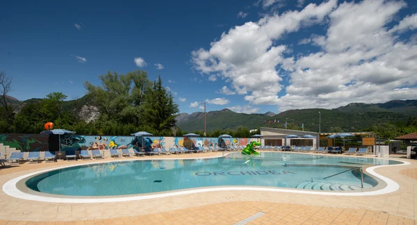 At Orchidea, a spacious, transparent swimming pool is encircled by deck chairs and sunshades, framed by a mountainous backdrop.