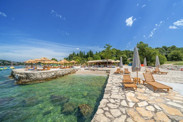 At Porton Nature Hideouts, wooden recliners with canopies are positioned on a stone terrace that overlooks pristine blue waters, encircled by lush vegetation.