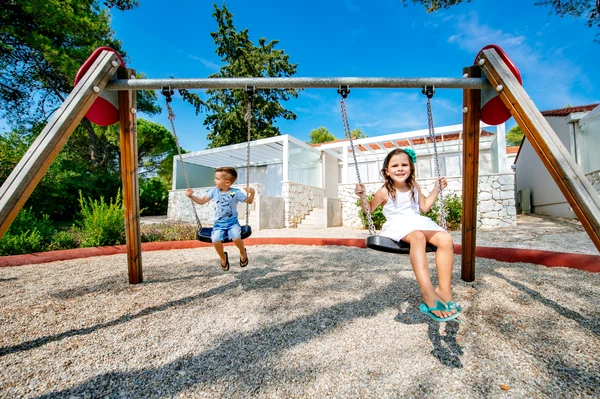At Zaton Holiday Resort, kids swing happily on a playset, encircled by gravel, vibrant greenery, and contemporary structures.