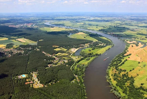 At Knaus Campingpark Bleckede/Elbtalaue, a river meanders through the landscape featuring woodlands, meadows, and dispersed structures, seen from an aerial perspective.