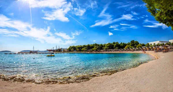 At Amadria Park Camping Sibenik, a golden sandy shore features sunbathers, a pedal boat glides on the crystal-clear water, encircled by vibrant green trees.