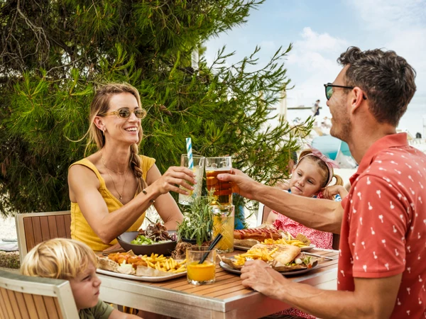 At Zaton Holiday Resort, a family raises their glasses at an outdoor table filled with food in a sunny, forested environment.