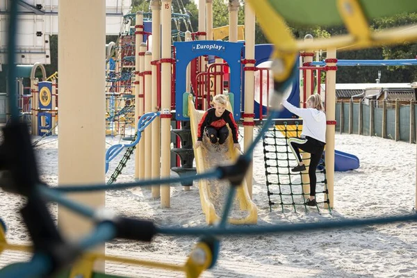 At Recreatieoord Wilhelm Tell, children are enjoying themselves on slides and climbing nets in a vibrant playground filled with sand, enclosed by fences and trees.