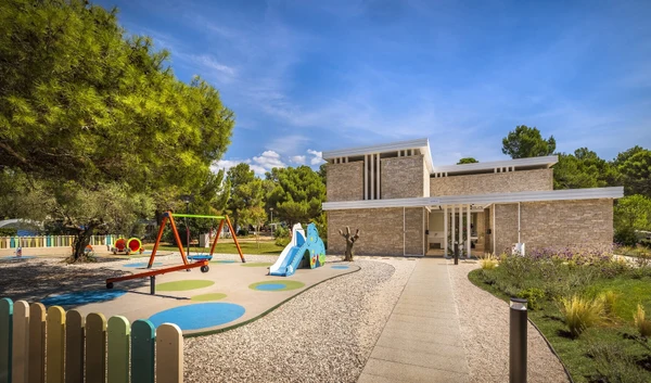 At Naturist Park Koversada, vibrant playground apparatus is being utilized in front of a contemporary stone structure surrounded by abundant greenery.