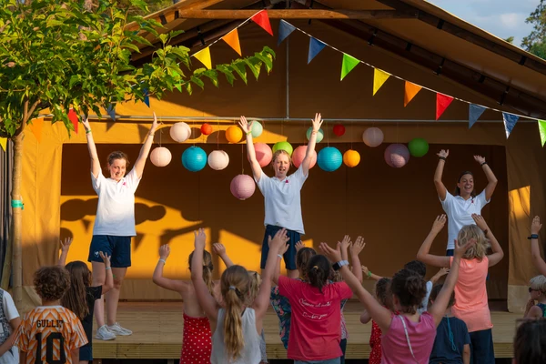 At Trasimeno Glamping Resort, instructors guide children in dance, on an outdoor stage adorned with vibrant lanterns and bunting, creating an energetic ambiance.