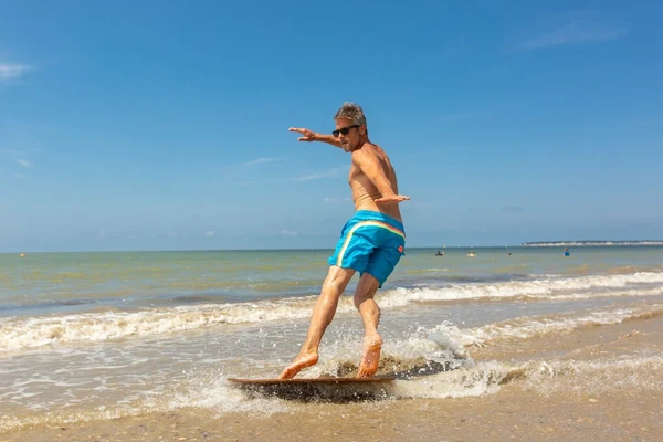 At Ondres Plage, an individual skimboards over shallow surf on a bright beach, keeping their balance and dressed in blue swim trunks.